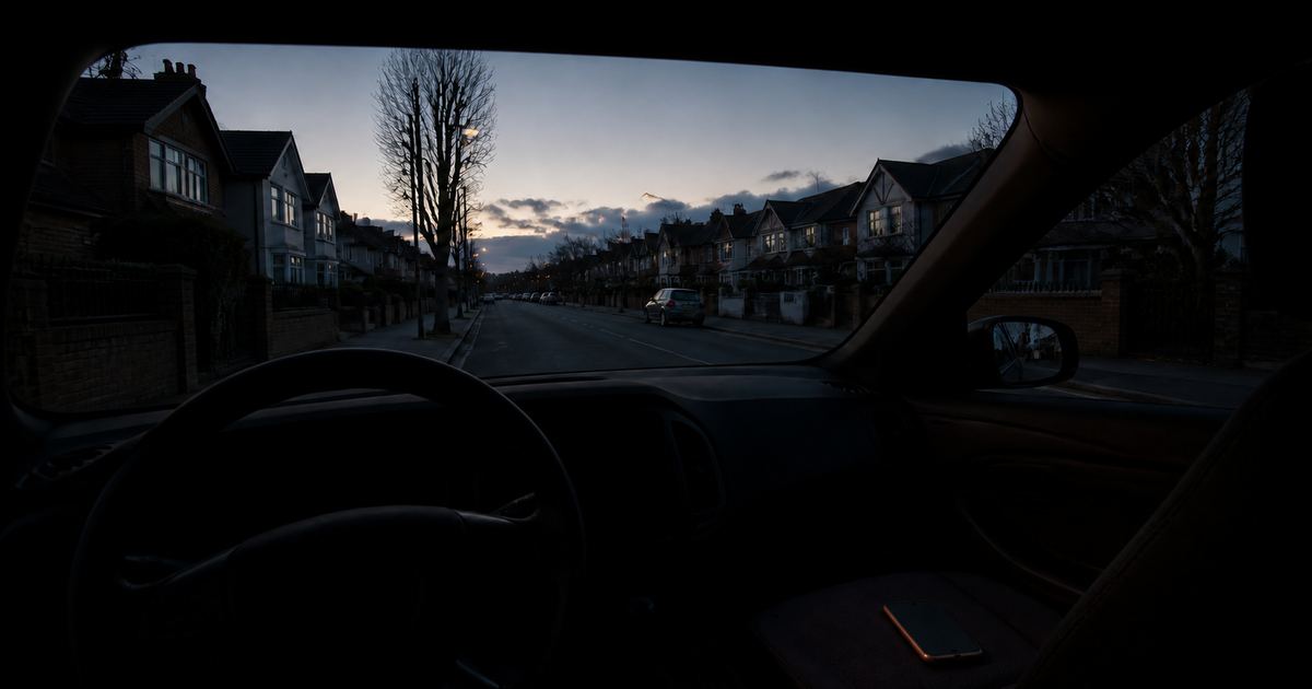 The dim interior of a parked car at dusk with an unoccupied steering wheel in silhouette and a phone on the passenger seat, a quiet street sharp through the windscreen